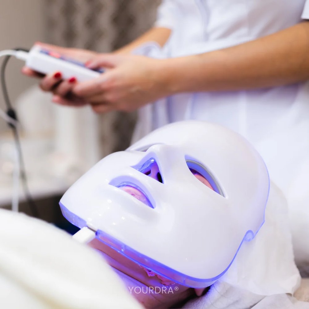 A client relaxing under a glowing LED face mask while a practitioner adjusts the controller